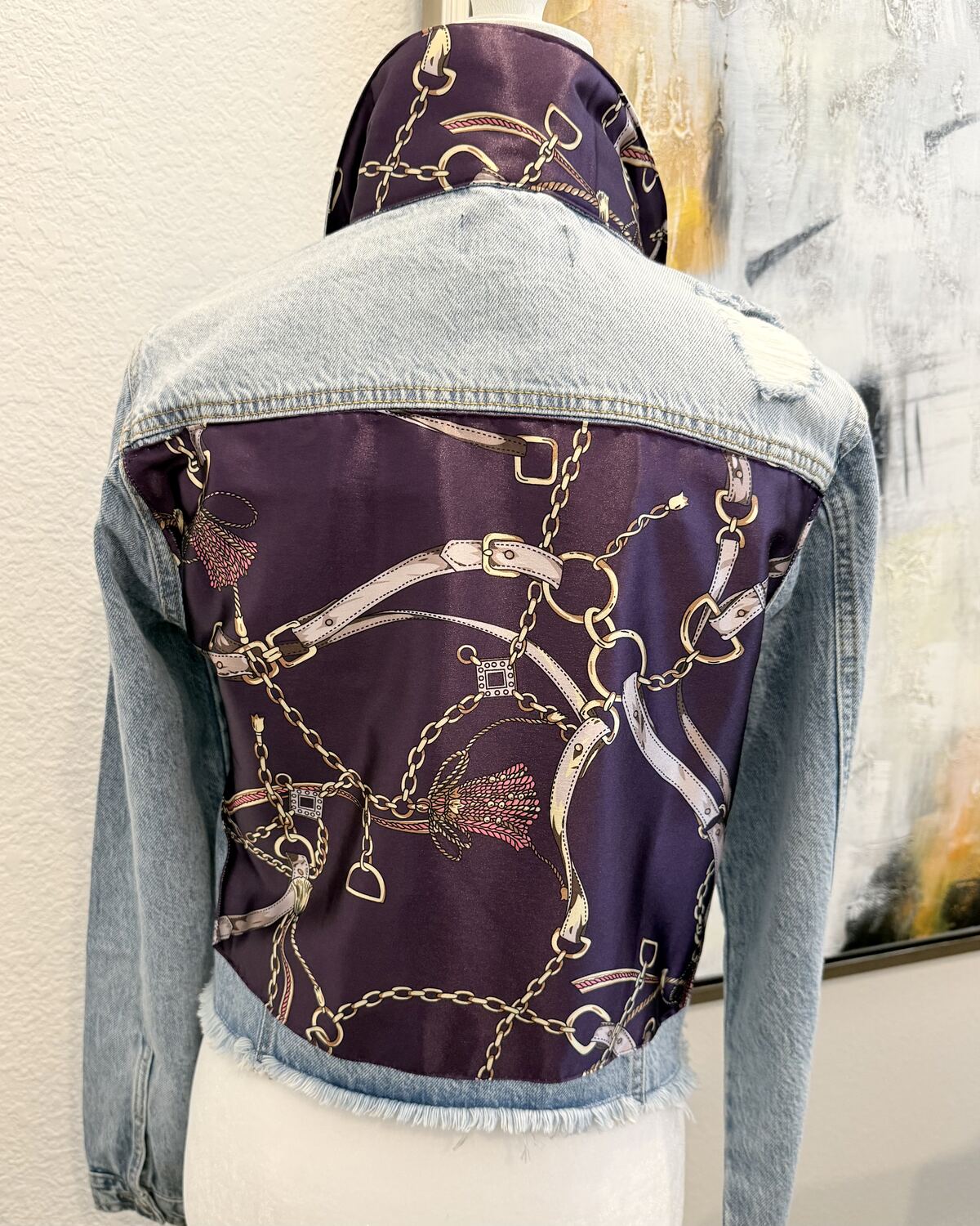 Back of a cropped, frayed-hem light denim jacket on a mannequin, the back panel backed with a purple and navy silk scarf print of gold chains and belts.
