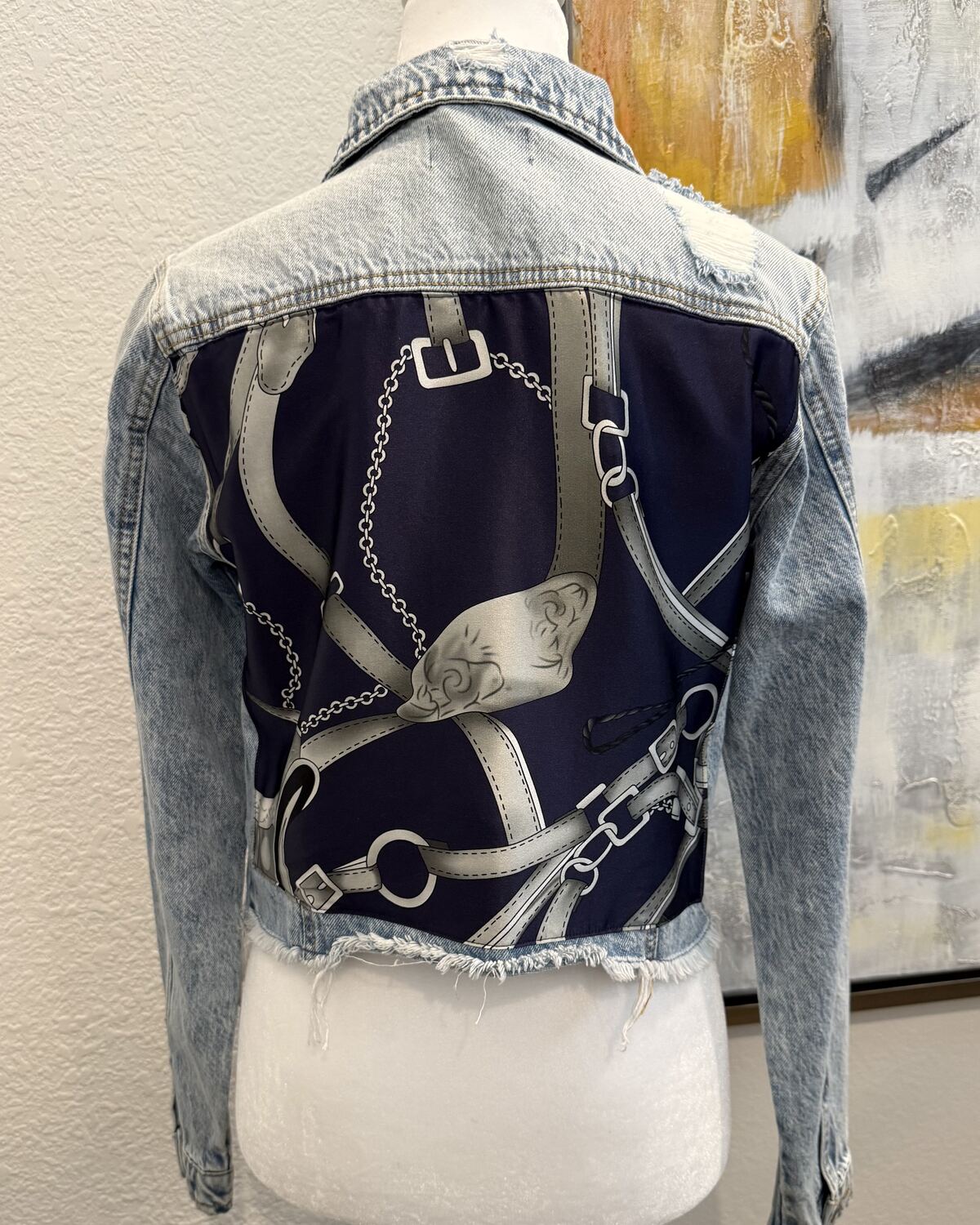 Back of a cropped, frayed-hem acid-wash denim jacket on a mannequin, the back panel backed with a navy silk scarf print of chains and buckled straps.