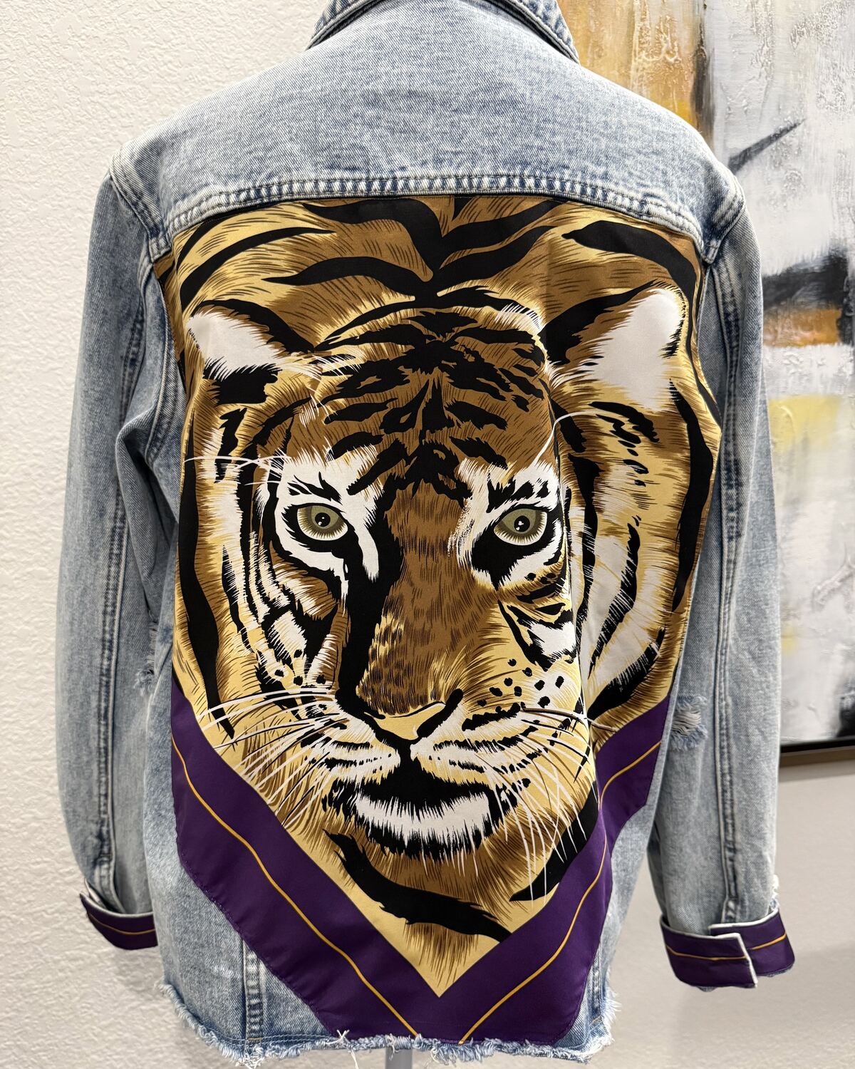 Back of a light denim jacket on a mannequin, the back panel backed with a gold tiger face on a deep purple border.