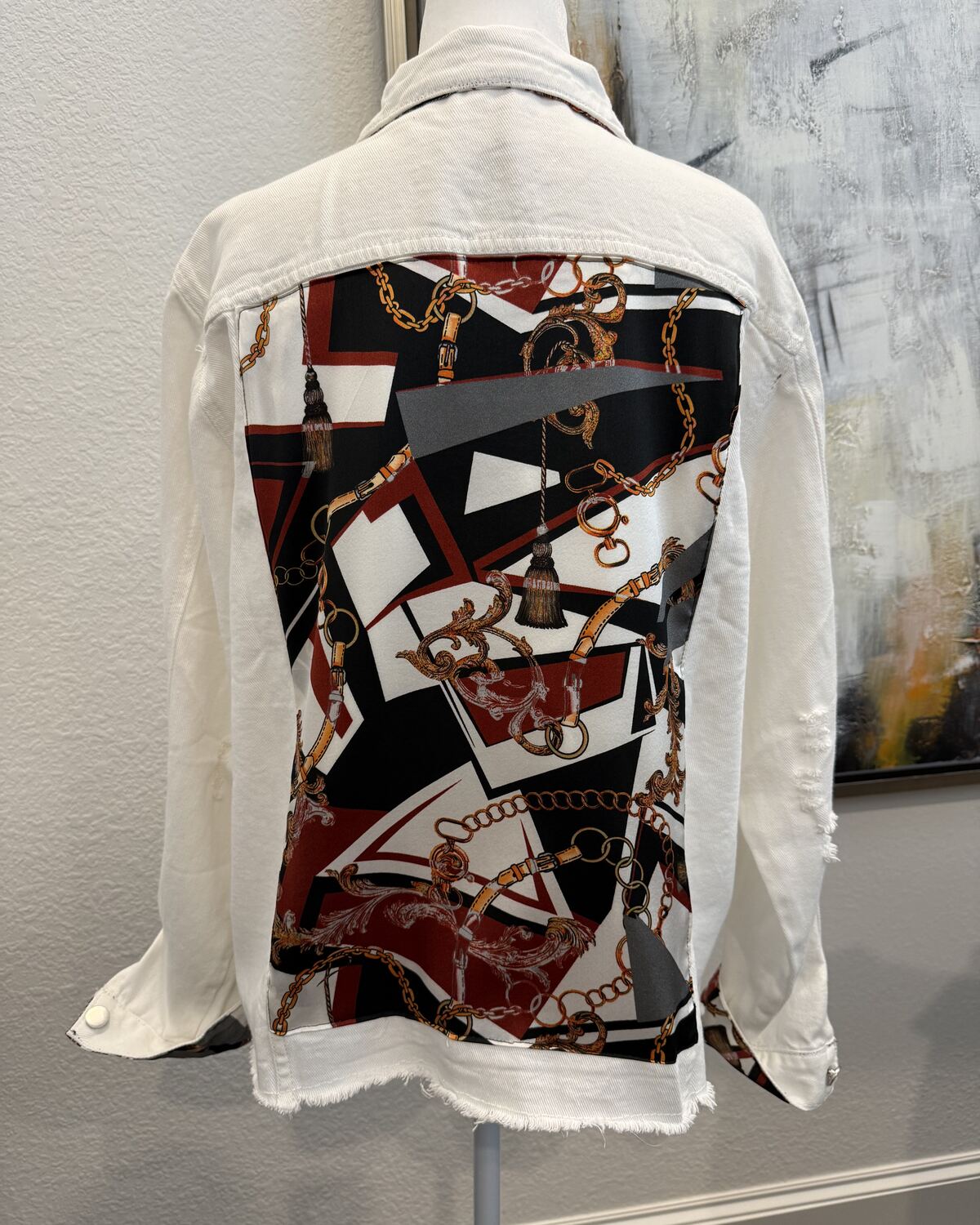 Back of a white denim jacket on a mannequin, the back panel backed with a bold geometric print in black, deep red, white, and gold chains.