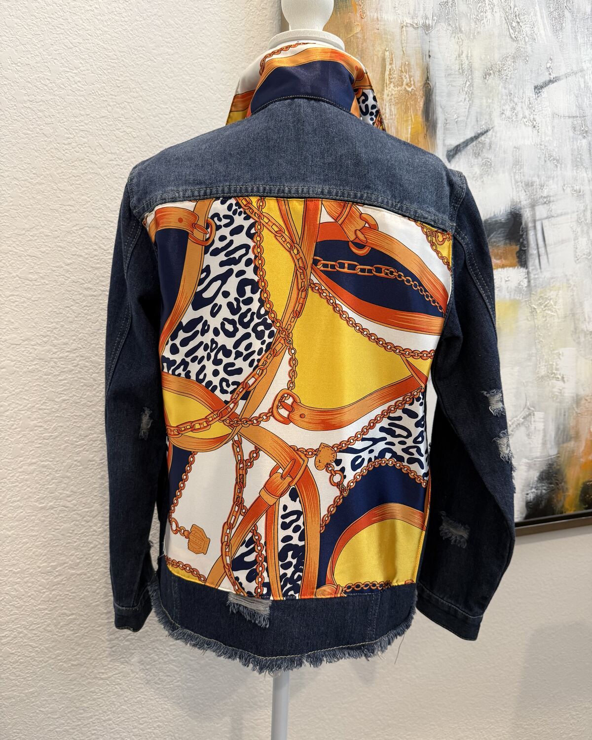 Back of a dark indigo denim jacket on a mannequin, the back panel backed with an orange, yellow, and leopard-spot scarf print with gold chains.